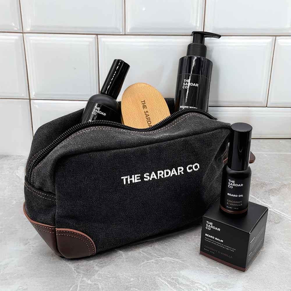 Black toiletry bag with 'The Sardar Co' branding, containing grooming products on a tiled bathroom floor.